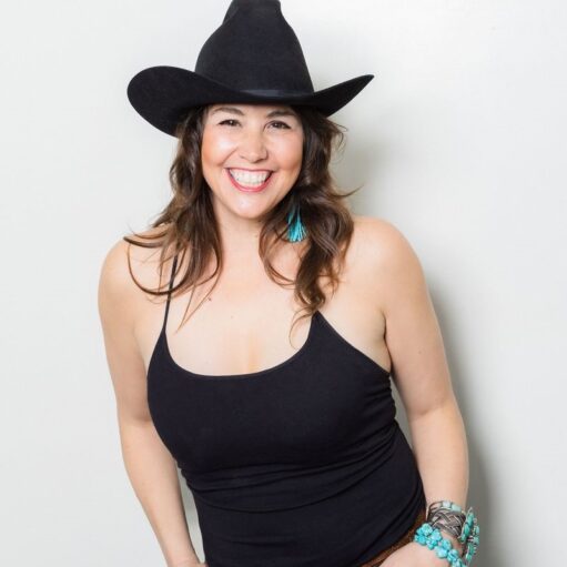 A smiling woman with wavy brown hair wearing a black cowboy hat and a black tank top. She is accessorized with turquoise tassel earrings and several chunky bracelets, including turquoise beads and a silver braided cuff, set against a plain white background.