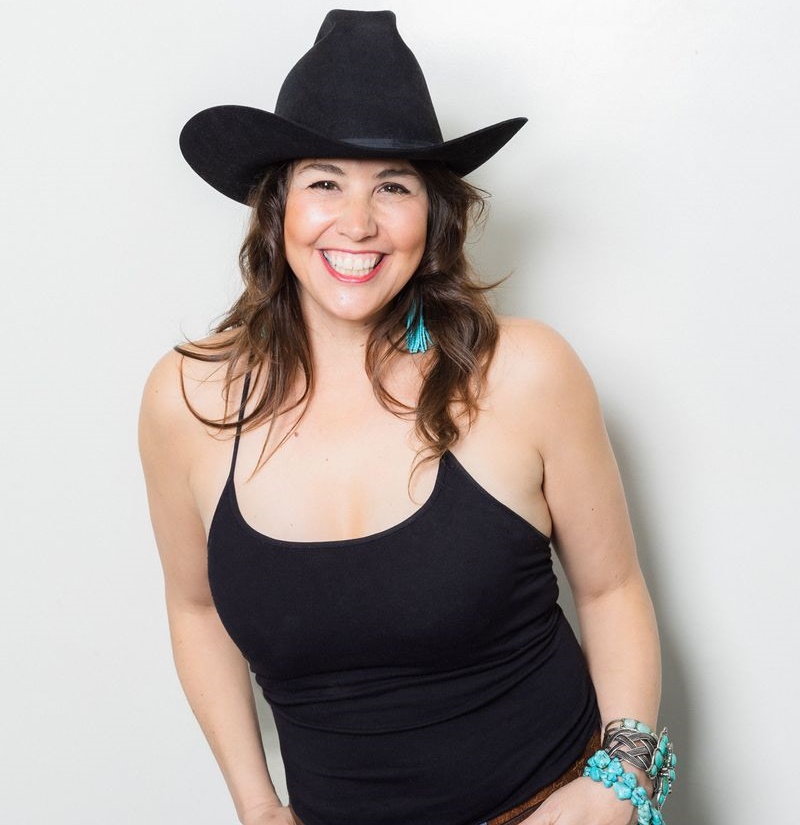 A smiling woman with wavy brown hair wearing a black cowboy hat and a black tank top. She is accessorized with turquoise tassel earrings and several chunky bracelets, including turquoise beads and a silver braided cuff, set against a plain white background.