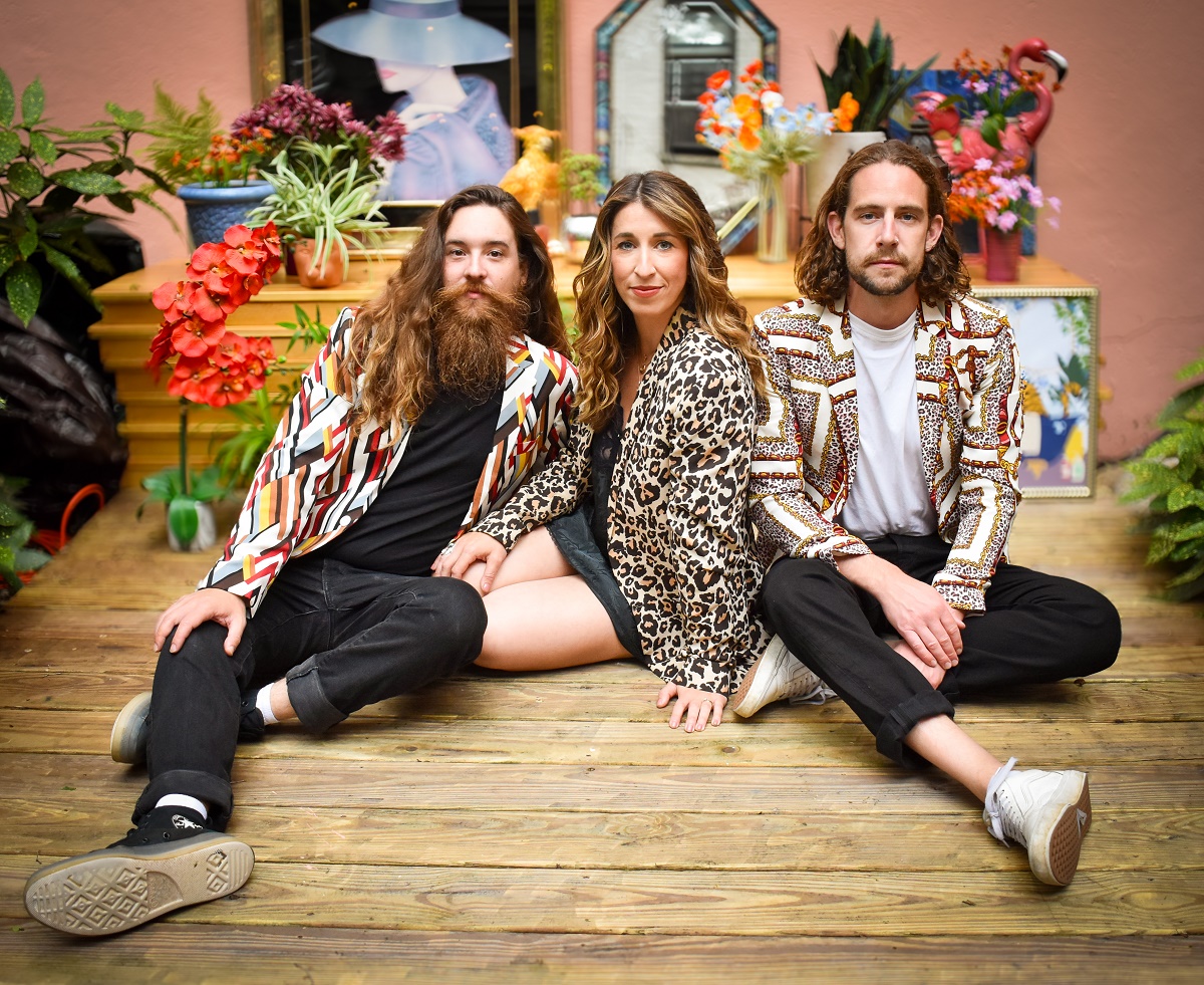 Three people sit on a wooden deck in front of a backdrop filled with colorful plants and eclectic decor. A man with a long beard and a geometric-patterned blazer sits on the left; a woman in a leopard-print blazer sits in the center; and a man with shoulder-length hair wearing a chain-link patterned blazer sits on the right.