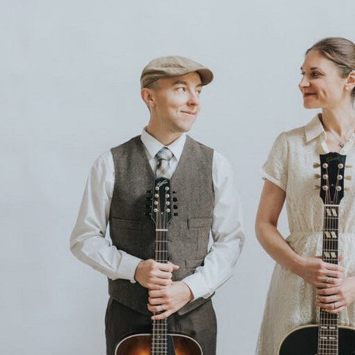 A man and a woman stand against a plain white background, looking toward one another with slight smiles. The man, on the left, wears a flat cap, a white dress shirt with a tie, and a brown waistcoat while holding a mandolin. The woman, on the right, wears an off-white lace dress and has her hair in a bun while holding an acoustic guitar.