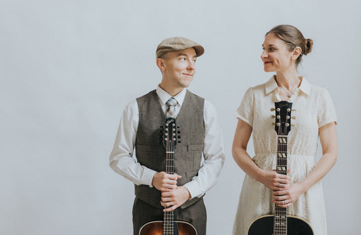 A man and a woman stand against a plain white background, looking toward one another with slight smiles. The man, on the left, wears a flat cap, a white dress shirt with a tie, and a brown waistcoat while holding a mandolin. The woman, on the right, wears an off-white lace dress and has her hair in a bun while holding an acoustic guitar.