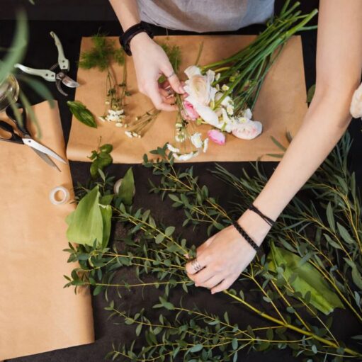 An overhead view of a florist’s hands arranging a bouquet on a dark workspace covered with brown craft paper. The scene includes various green stems, delicate white and pink flowers, and floristry tools like scissors and shears.
