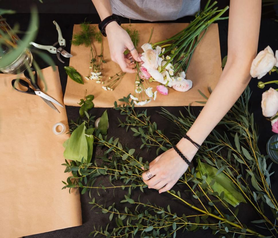 An overhead view of a florist’s hands arranging a bouquet on a dark workspace covered with brown craft paper. The scene includes various green stems, delicate white and pink flowers, and floristry tools like scissors and shears.