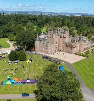 An aerial view of a large, historic stone castle surrounded by lush green lawns. People are gathered on the grass for an outdoor event featuring white tents and a large, colourful inflatable obstacle course.