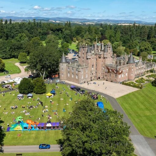 An aerial view of a large, historic stone castle surrounded by lush green lawns. People are gathered on the grass for an outdoor event featuring white tents and a large, colourful inflatable obstacle course.