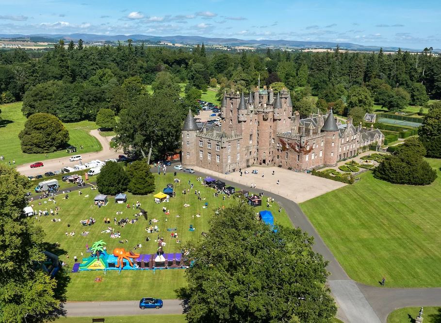 An aerial view of a large, historic stone castle surrounded by lush green lawns. People are gathered on the grass for an outdoor event featuring white tents and a large, colourful inflatable obstacle course.