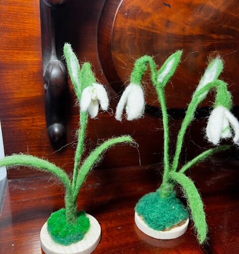 Two needle-felted snowdrop flowers stand on small, circular wooden bases against a dark wood background. The delicate sculptures feature slender green stems and leaves with drooping white bell-shaped petals, mimicking the appearance of real spring blooms.
