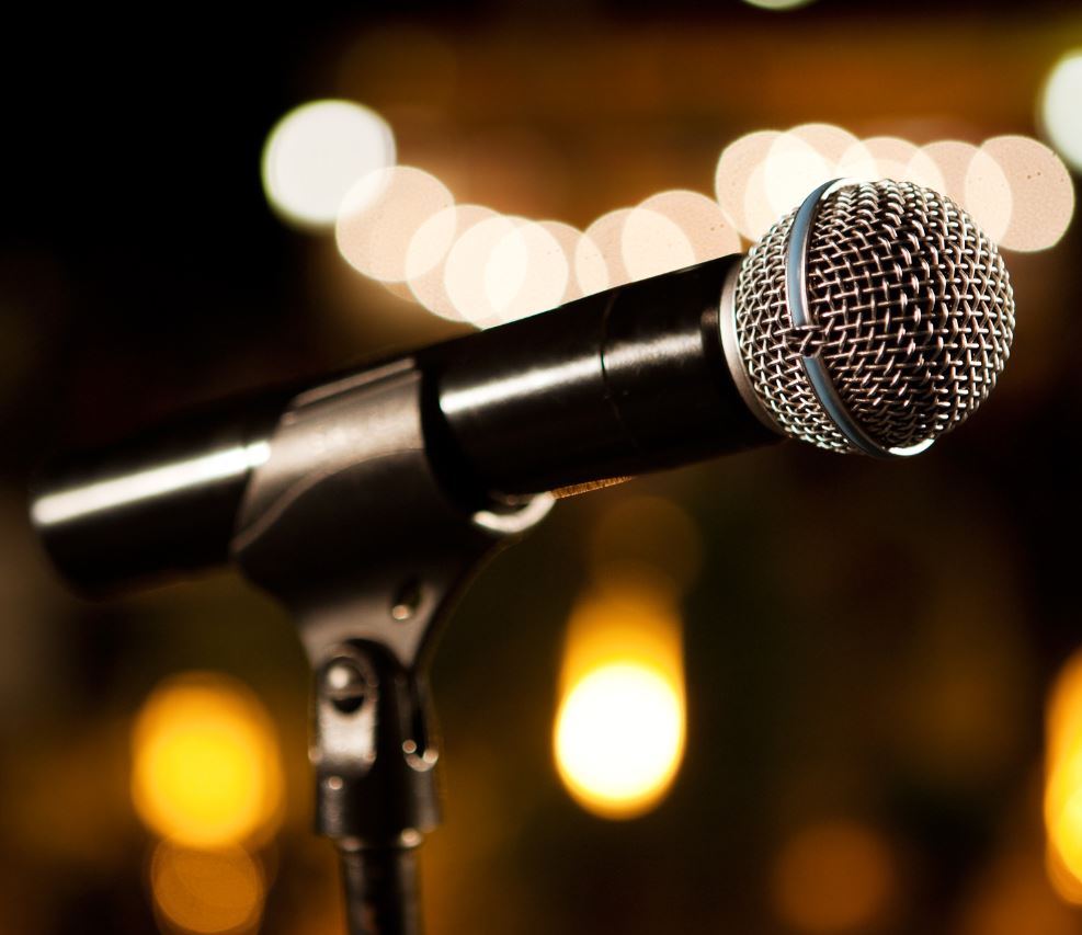 A close-up, angled shot of a black dynamic microphone in a stand. The background is dark and out of focus, featuring warm, golden bokeh lights that suggest a stage or event setting.