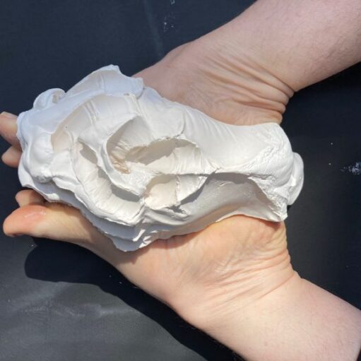 An overhead shot of two hands cupped together, holding a large, irregular lump of white clay or plaster. The material has a rough, sculpted texture with deep indentations and folds. The background is a flat, dark grey surface.