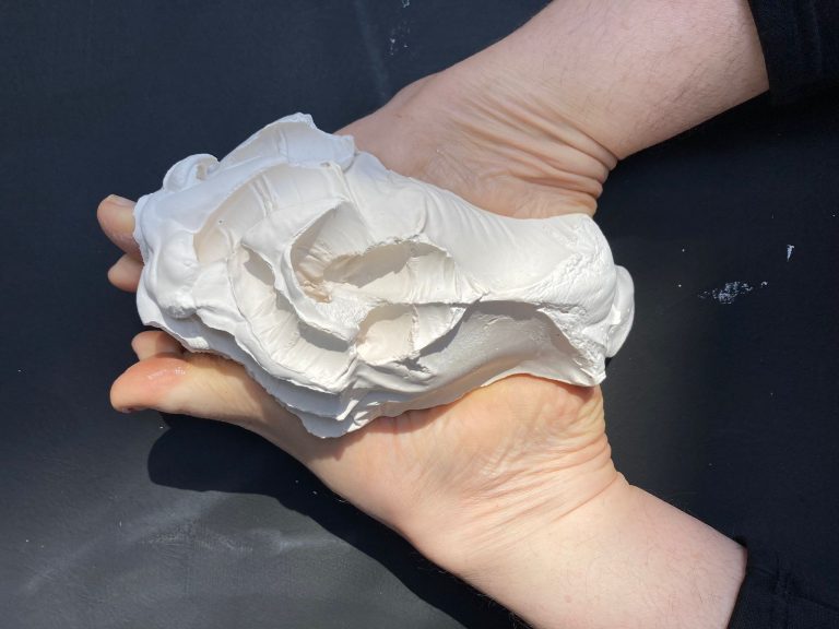An overhead shot of two hands cupped together, holding a large, irregular lump of white clay or plaster. The material has a rough, sculpted texture with deep indentations and folds. The background is a flat, dark grey surface.
