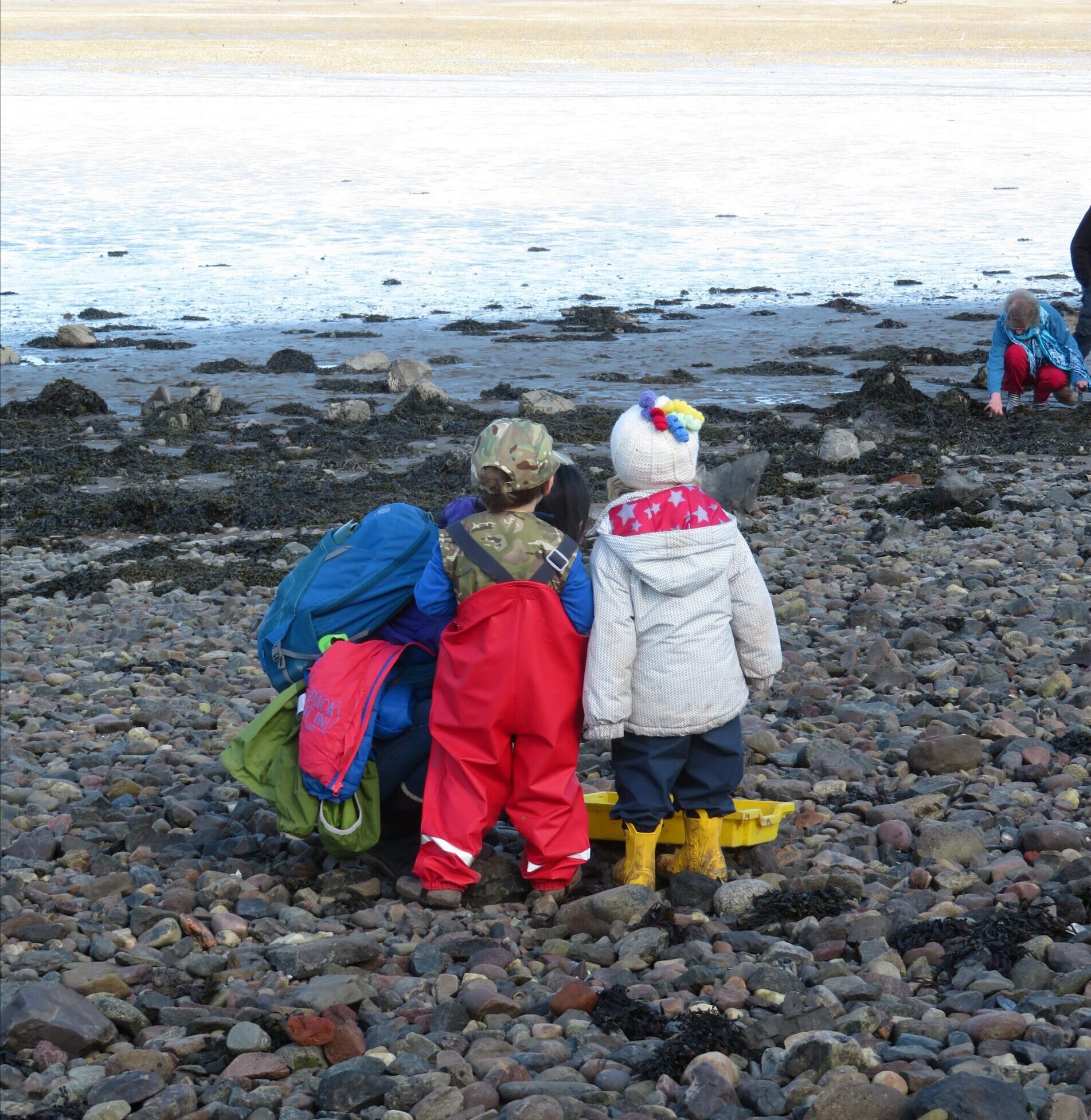 Two young children, seen from behind, stand on a rocky beach by the water's edge. One child wears bright red waterproof overalls and a camouflage hat, while the other wears a white jacket, dark pants, yellow boots, and a white knit hat with colorful pom-poms. A person with a blue backpack stands next to them, and in the background, another person is crouched down near the tide line.