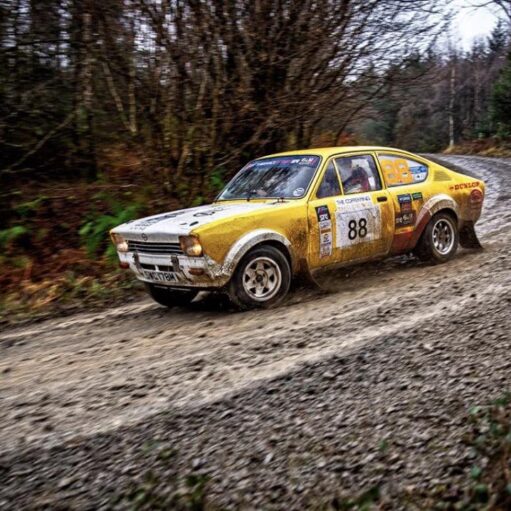 A yellow and white vintage Opel Kadett rally car, number 88, speeds along a muddy gravel track through a dense forest. The car is splattered with mud, capturing a sense of motion on a sharp bend.