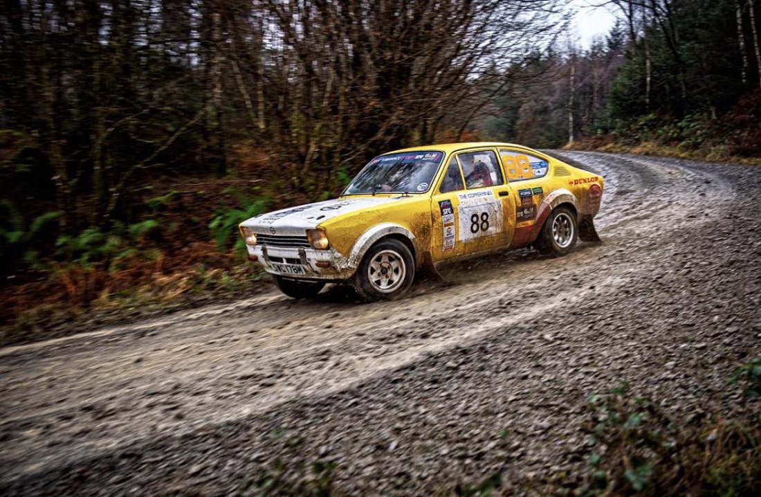 A yellow and white vintage Opel Kadett rally car, number 88, speeds along a muddy gravel track through a dense forest. The car is splattered with mud, capturing a sense of motion on a sharp bend.