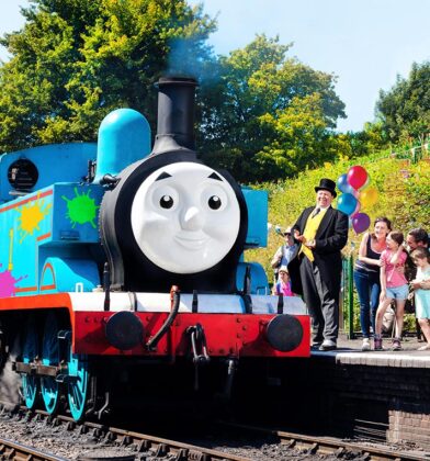 A life-sized Thomas the Tank Engine steam locomotive, decorated with colorful paint splats, sits at a train station platform. Standing on the platform next to the train is a man dressed as Sir Topham Hatt (The Fat Controller) in a top hat and morning suit, gesturing toward a group of excited children and adults. Colorful balloons and lush green trees are visible in the background under a bright, sunny sky.