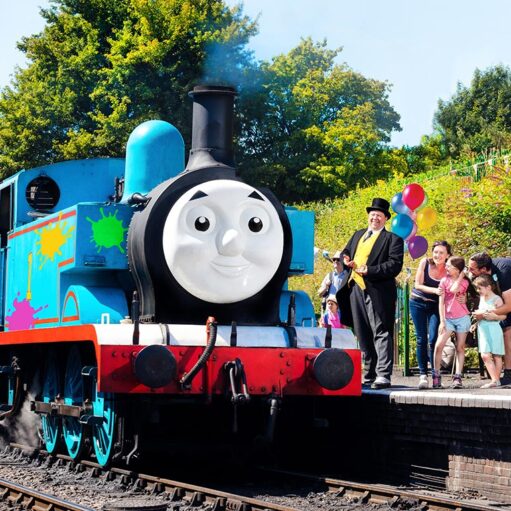 A life-sized Thomas the Tank Engine steam locomotive, decorated with colorful paint splats, sits at a train station platform. Standing on the platform next to the train is a man dressed as Sir Topham Hatt (The Fat Controller) in a top hat and morning suit, gesturing toward a group of excited children and adults. Colorful balloons and lush green trees are visible in the background under a bright, sunny sky.