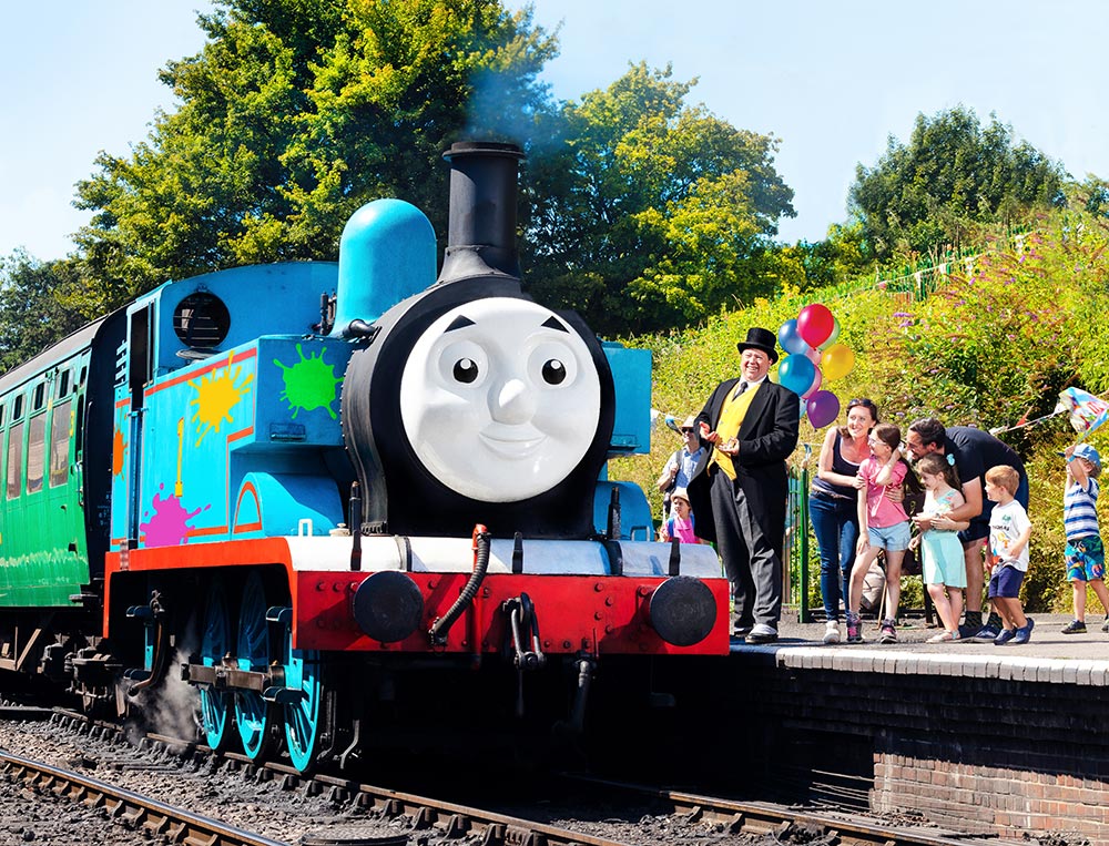 A life-sized Thomas the Tank Engine steam locomotive, decorated with colorful paint splats, sits at a train station platform. Standing on the platform next to the train is a man dressed as Sir Topham Hatt (The Fat Controller) in a top hat and morning suit, gesturing toward a group of excited children and adults. Colorful balloons and lush green trees are visible in the background under a bright, sunny sky.