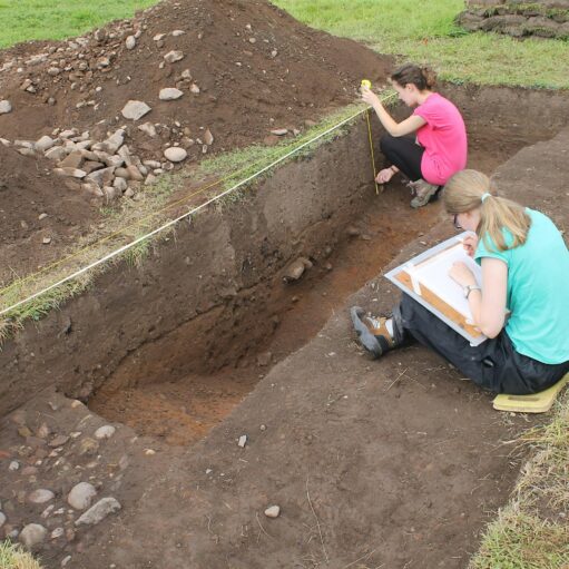 An archaeological excavation site showing two people working in a deep, rectangular trench cut into grassy ground. One person in a pink shirt is crouched down using a measuring tape against the trench wall, while another person in a turquoise shirt sits on the edge, sketching or taking notes on a clipboard. Large mounds of excavated soil and rocks are piled beside the trench.