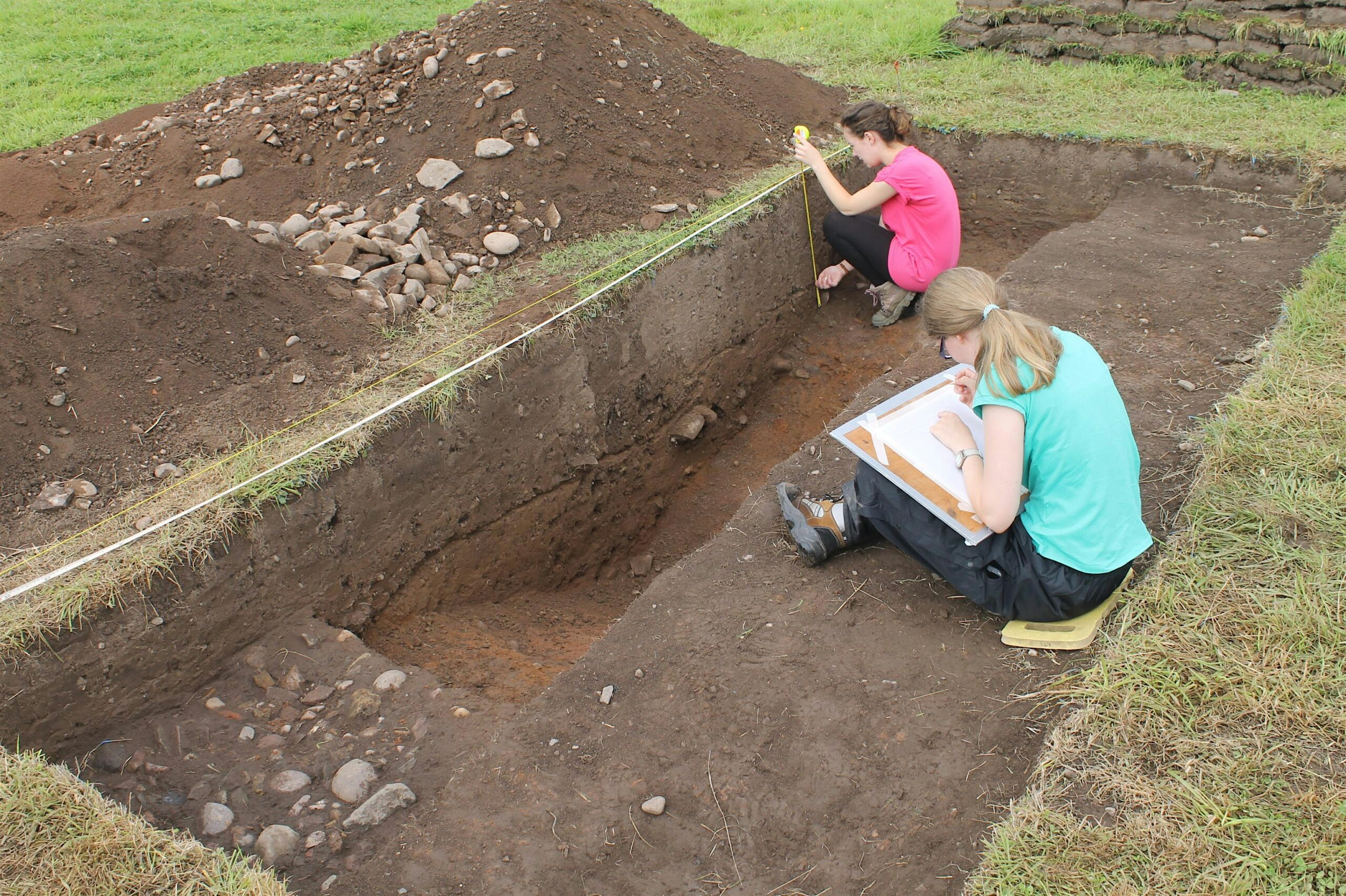 An archaeological excavation site showing two people working in a deep, rectangular trench cut into grassy ground. One person in a pink shirt is crouched down using a measuring tape against the trench wall, while another person in a turquoise shirt sits on the edge, sketching or taking notes on a clipboard. Large mounds of excavated soil and rocks are piled beside the trench.