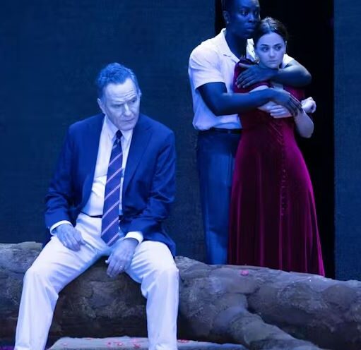 A dramatic stage production scene bathed in blue light. In the foreground, a man in a blue suit, white trousers, and a striped tie sits on a large, textured stone or log, looking off to the side with a serious expression. Behind him, another person in a white shirt and blue trousers stands with their arms wrapped around a woman in a long, dark red velvet dress, who looks concerned. The background is a dark, solid wall.