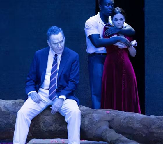 A dramatic stage production scene bathed in blue light. In the foreground, a man in a blue suit, white trousers, and a striped tie sits on a large, textured stone or log, looking off to the side with a serious expression. Behind him, another person in a white shirt and blue trousers stands with their arms wrapped around a woman in a long, dark red velvet dress, who looks concerned. The background is a dark, solid wall.