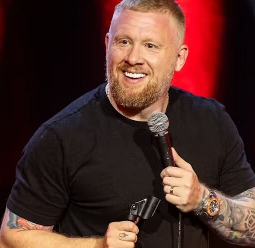 A man with short blonde hair and a reddish-blonde beard is smiling while holding a microphone on stage. He is wearing a black t-shirt and has visible tattoos on both arms. He is also wearing a watch with an orange bezel and a gold wedding band. The background consists of blurred red stage lighting.