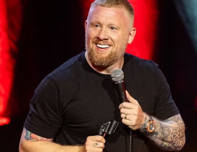 A man with short blonde hair and a reddish-blonde beard is smiling while holding a microphone on stage. He is wearing a black t-shirt and has visible tattoos on both arms. He is also wearing a watch with an orange bezel and a gold wedding band. The background consists of blurred red stage lighting.