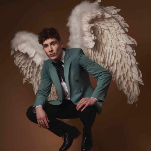 A studio portrait of a man with dark curly hair posing against a warm brown background. He is wearing a teal blazer, a white shirt with a black bow tie, and black trousers while sporting a large pair of white feathered angel wings. He is crouched in a mid-air jumping position, looking directly at the camera with a neutral expression.