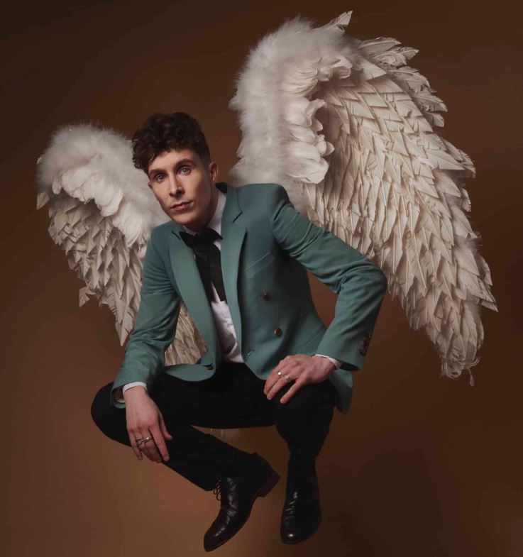 A studio portrait of a man with dark curly hair posing against a warm brown background. He is wearing a teal blazer, a white shirt with a black bow tie, and black trousers while sporting a large pair of white feathered angel wings. He is crouched in a mid-air jumping position, looking directly at the camera with a neutral expression.