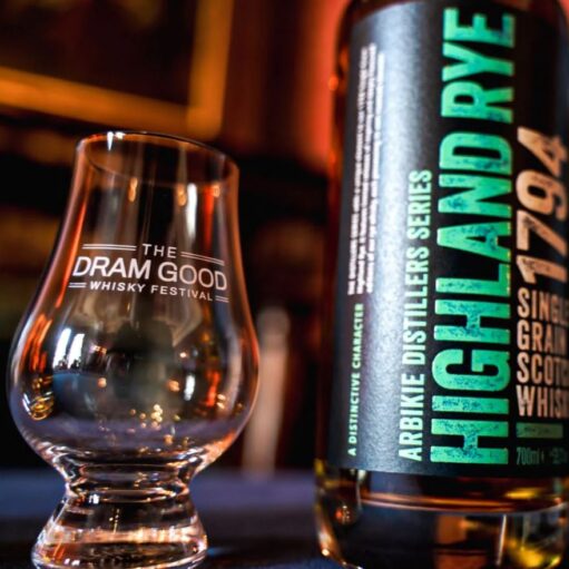A close-up of a bottle of Arbikie Highland Rye 1794 Single Grain Scotch Whisky standing next to a Glencairn tasting glass etched with "The Dram Good Whisky Festival" logo. The background is dimly lit with warm, ambient tones.