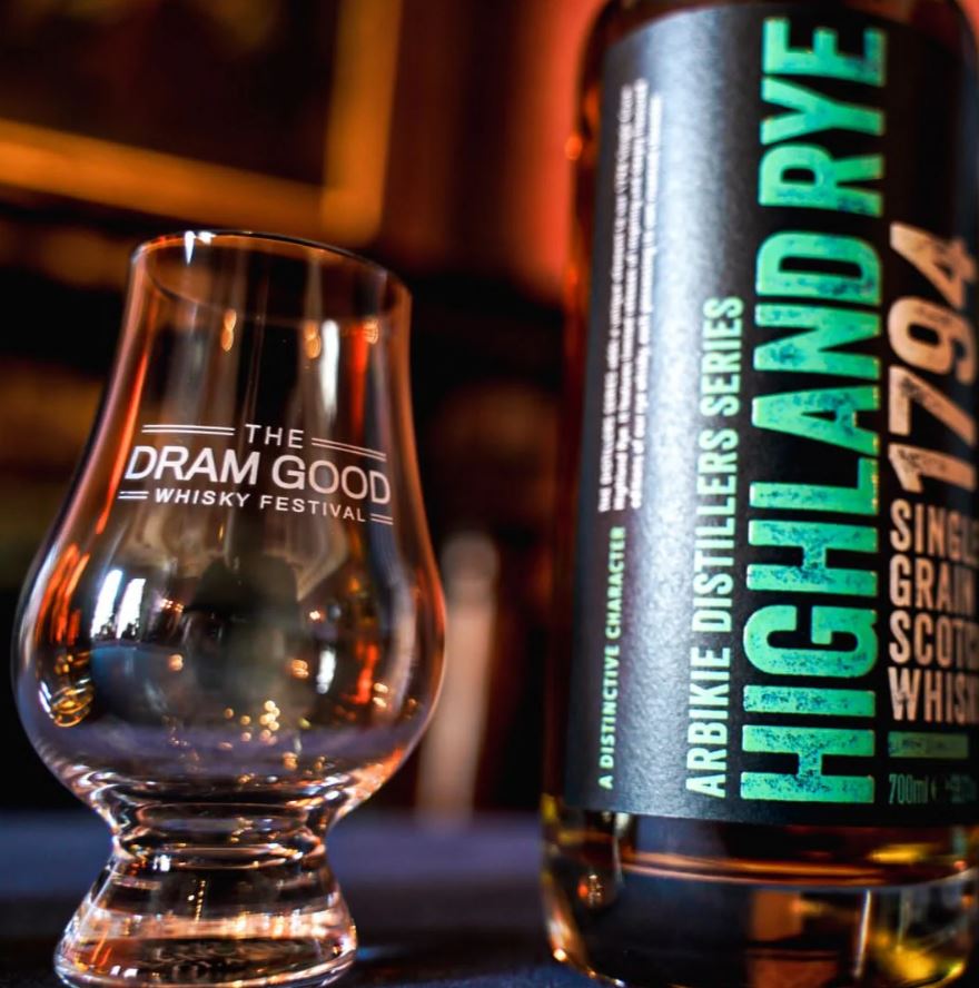 A close-up of a bottle of Arbikie Highland Rye 1794 Single Grain Scotch Whisky standing next to a Glencairn tasting glass etched with "The Dram Good Whisky Festival" logo. The background is dimly lit with warm, ambient tones.