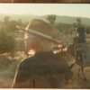 Vintage-style photograph of a person wearing a wide-brimmed hat standing beside a film camera in a desert landscape, with scrub vegetation, distant mountains and warm golden sunlight creating a cinematic atmosphere.