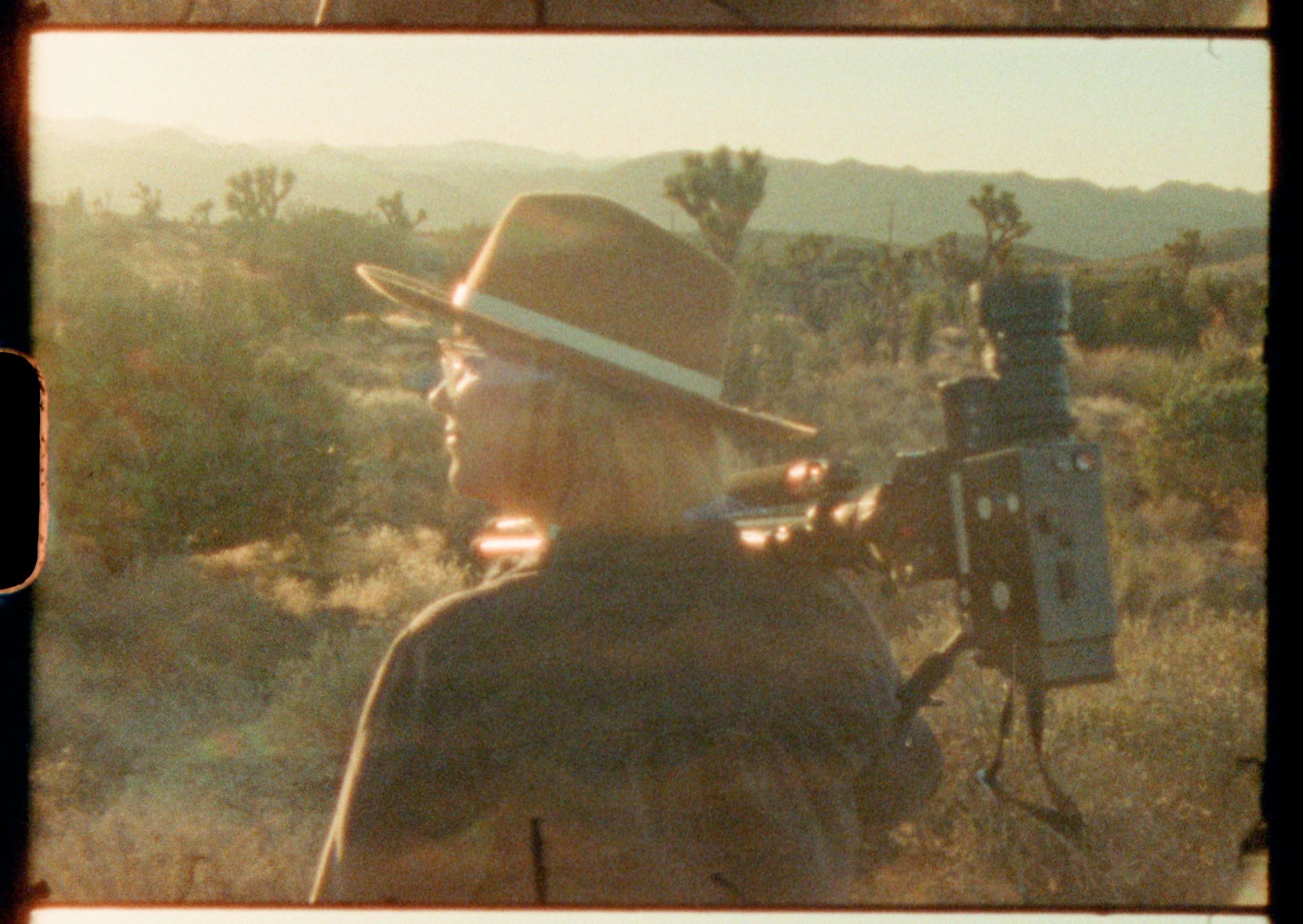 Vintage-style photograph of a person wearing a wide-brimmed hat standing beside a film camera in a desert landscape, with scrub vegetation, distant mountains and warm golden sunlight creating a cinematic atmosphere.