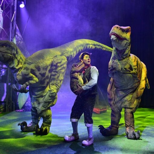 A performer in a vest and clown shoes stands under purple stage lights, cradling a small dinosaur puppet while flanked by two large, realistic dinosaur costumes on a green-lit stage.