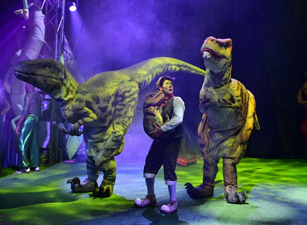 A performer in a vest and clown shoes stands under purple stage lights, cradling a small dinosaur puppet while flanked by two large, realistic dinosaur costumes on a green-lit stage.