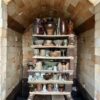 A view inside a brick kiln with an arched ceiling, containing several wooden shelves stacked with various handmade pottery. The shelves are filled with ceramic items including vases, bowls, and mugs in earth tones like brown, tan, and grey.