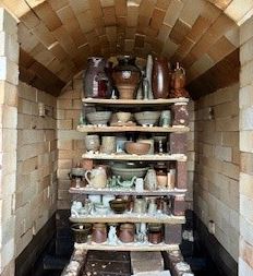 A view inside a brick kiln with an arched ceiling, containing several wooden shelves stacked with various handmade pottery. The shelves are filled with ceramic items including vases, bowls, and mugs in earth tones like brown, tan, and grey.