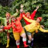 Four people are posing joyfully outdoors in front of a dense backdrop of large green leaves. They are dressed in a vibrant array of colorful rain jackets (green, orange, red, and yellow), matching beanies, striped scarves, shorts, and bright rubber boots. Each person has their arms raised and wide smiles on their faces, conveying a sense of energetic fun.