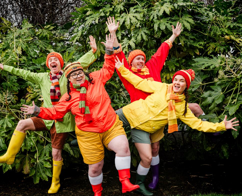 Four people are posing joyfully outdoors in front of a dense backdrop of large green leaves. They are dressed in a vibrant array of colorful rain jackets (green, orange, red, and yellow), matching beanies, striped scarves, shorts, and bright rubber boots. Each person has their arms raised and wide smiles on their faces, conveying a sense of energetic fun.