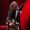 A long-haired musician in a black t-shirt sings into a microphone while playing a white electric guitar on stage. The background is washed in dramatic red lighting, emphasizing the high-energy atmosphere of a live performance.