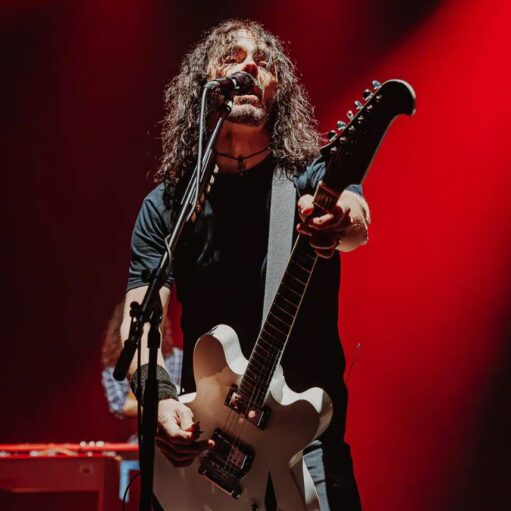 A long-haired musician in a black t-shirt sings into a microphone while playing a white electric guitar on stage. The background is washed in dramatic red lighting, emphasizing the high-energy atmosphere of a live performance.