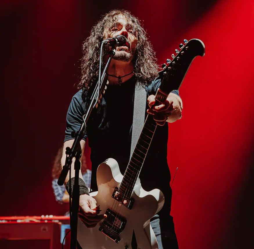 A long-haired musician in a black t-shirt sings into a microphone while playing a white electric guitar on stage. The background is washed in dramatic red lighting, emphasizing the high-energy atmosphere of a live performance.