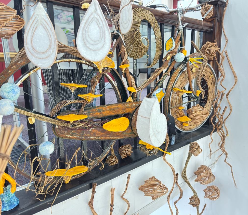 A vintage, rusted bicycle is mounted as a mixed-media art installation against a black metal railing. The bike is intricately decorated with woven wicker elements, large white tear-drop shapes, and bright yellow fungal-like protrusions that appear to be growing from the frame and wheels. Other abstract woven shapes and light blue spheres are suspended around the bicycle, creating a whimsical, organic display.