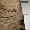 A close-up of an ancient, weathered stone slab featuring intricate carvings. The visible designs include swirling Celtic-style patterns and wavy horizontal lines etched into the rough, tan-colored surface. In the background, a blurred informational museum sign is partially visible on a white wall.
