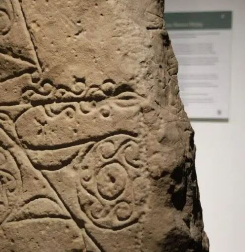 A close-up of an ancient, weathered stone slab featuring intricate carvings. The visible designs include swirling Celtic-style patterns and wavy horizontal lines etched into the rough, tan-colored surface. In the background, a blurred informational museum sign is partially visible on a white wall.