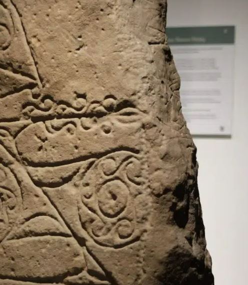A close-up of an ancient, weathered stone slab featuring intricate carvings. The visible designs include swirling Celtic-style patterns and wavy horizontal lines etched into the rough, tan-colored surface. In the background, a blurred informational museum sign is partially visible on a white wall.