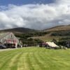 A wide-angle outdoor shot of a vibrant green lawn leading toward a rustic barn and several event tents. People are scattered throughout the area under a dynamic, cloudy sky, with rolling hills and a dense treeline providing a scenic backdrop in the distance.