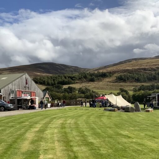 A wide-angle outdoor shot of a vibrant green lawn leading toward a rustic barn and several event tents. People are scattered throughout the area under a dynamic, cloudy sky, with rolling hills and a dense treeline providing a scenic backdrop in the distance.