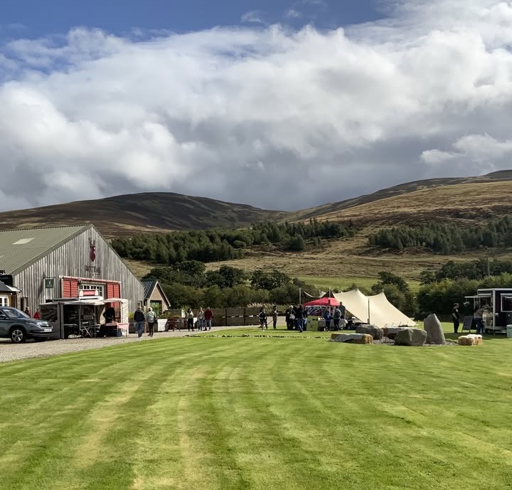 A wide-angle outdoor shot of a vibrant green lawn leading toward a rustic barn and several event tents. People are scattered throughout the area under a dynamic, cloudy sky, with rolling hills and a dense treeline providing a scenic backdrop in the distance.