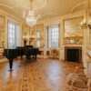 An opulent hall featuring intricate parquet wood flooring and walls adorned with large, gold-framed mirrors and elegant wall sconces. A black grand piano sits near tall windows, while a crystal chandelier hangs from the ornate white ceiling. The room also includes a marble fireplace and classic upholstered chairs.