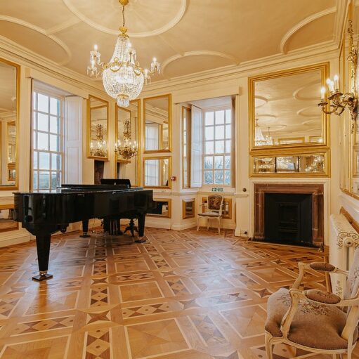 An opulent hall featuring intricate parquet wood flooring and walls adorned with large, gold-framed mirrors and elegant wall sconces. A black grand piano sits near tall windows, while a crystal chandelier hangs from the ornate white ceiling. The room also includes a marble fireplace and classic upholstered chairs.